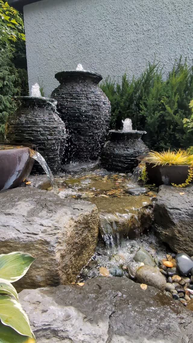 Three dark textured urn fountains pouring water into a rocky stream surrounded by greenery.