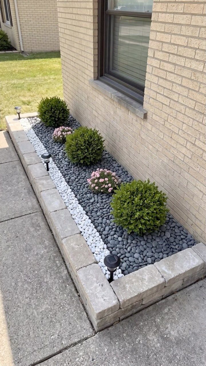 Green evergreen shrubs and pink flowers planted in a stone-bordered garden bed with grey pebbles.