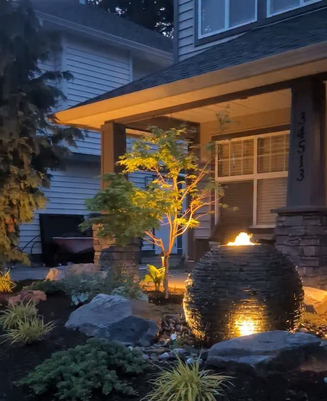 A lit stone water feature sits among rocks and small plants in a landscaped front yard.