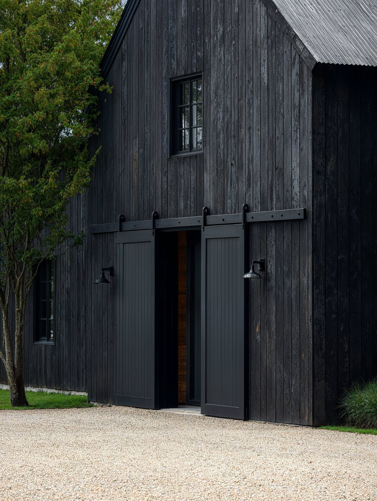 9 Modern Farmhouse Shed Ideas for 2026 3 Black wooden barn style shed with sliding doors and a gravel foreground.