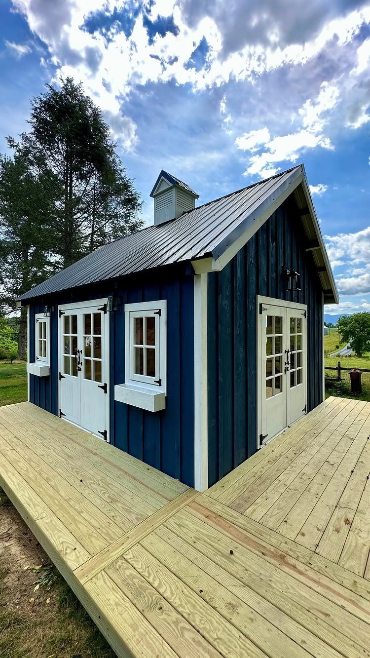 9 Modern Farmhouse Shed Ideas for 2026 9 A blue modern farmhouse style shed with white trim and a metal roof on a wooden deck.