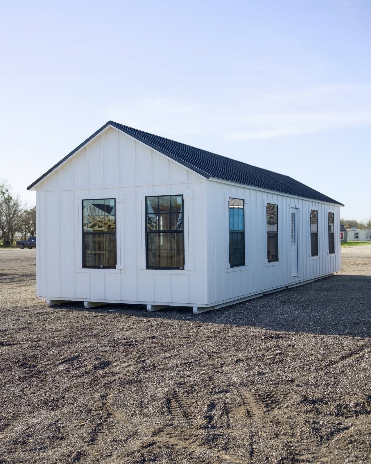 9 Modern Farmhouse Shed Ideas for 2026 1 A long white board and batten shed with black windows on a gravel lot.