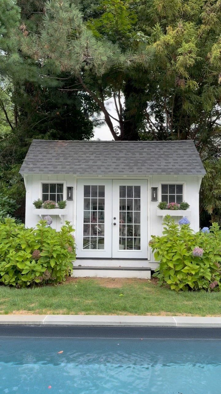 9 Modern Farmhouse Shed Ideas for 2026 5 A white garden shed with French doors and flower boxes sits on a green lawn.