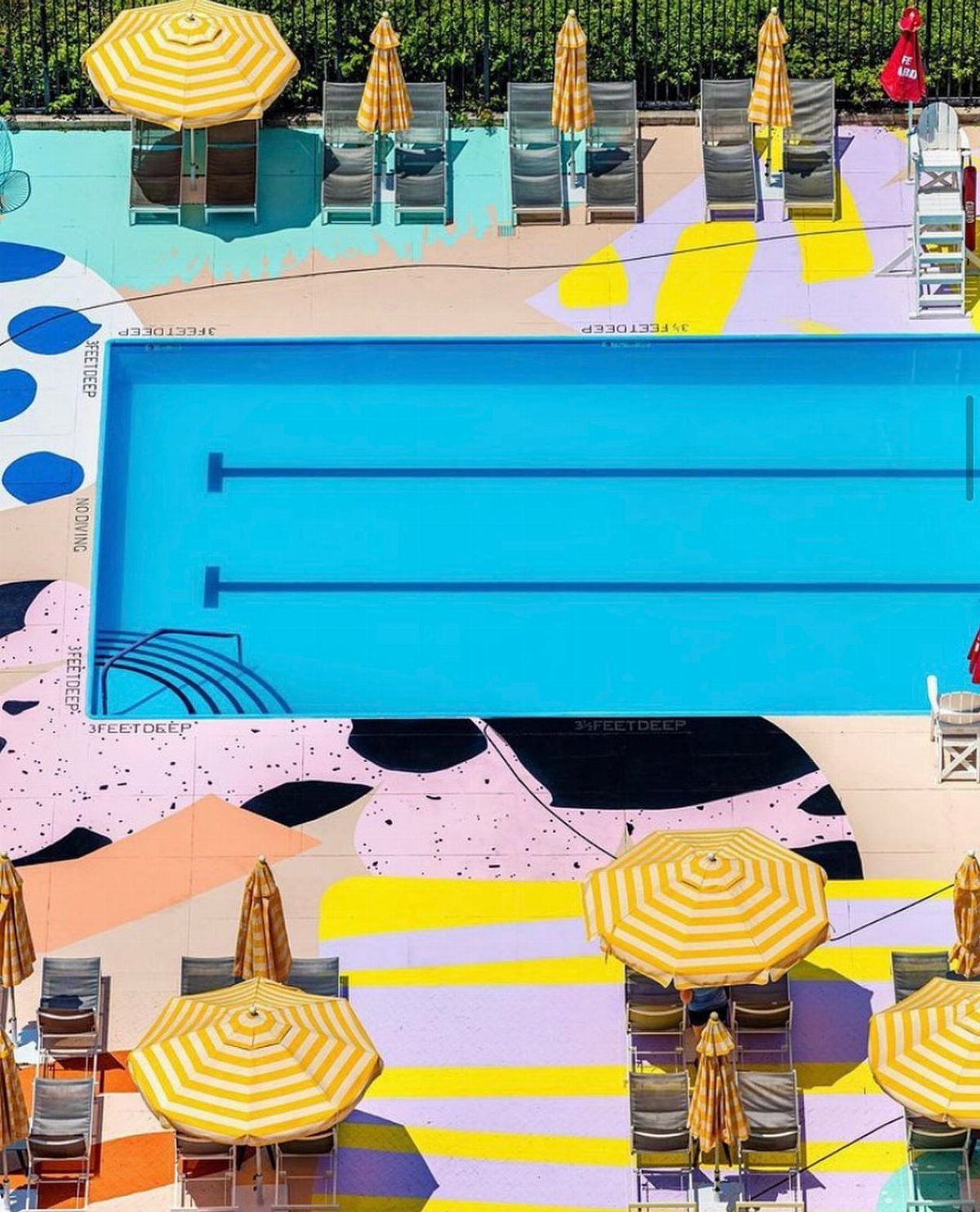 Pool Deck Ideas: 20 Designs From Breeze Blocks to Mountain Views 10 Aerial view of a swimming pool with a brightly painted abstract pattern deck and striped umbrellas.