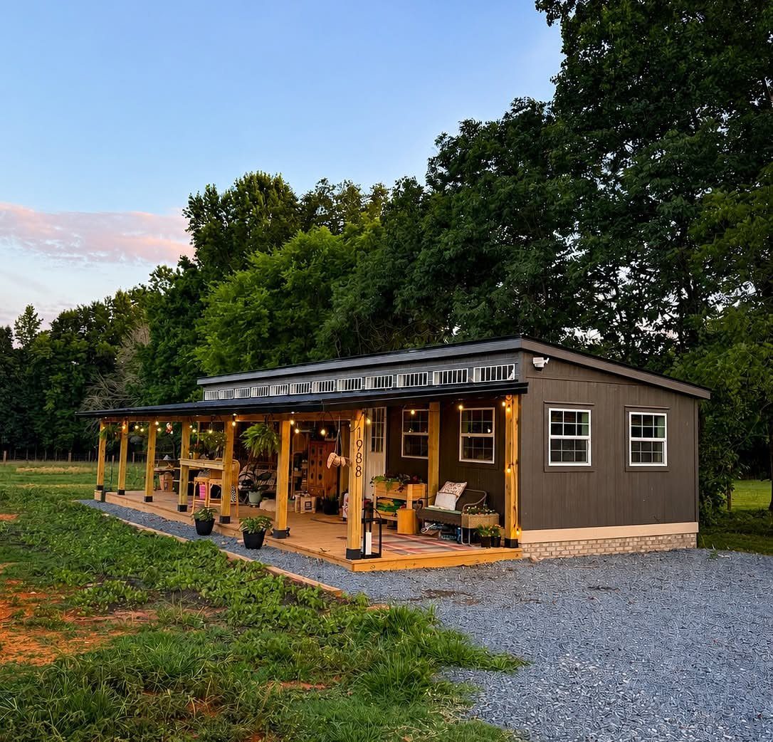 A dark grey converted shed house with a wooden porch and string lights in a grassy field.