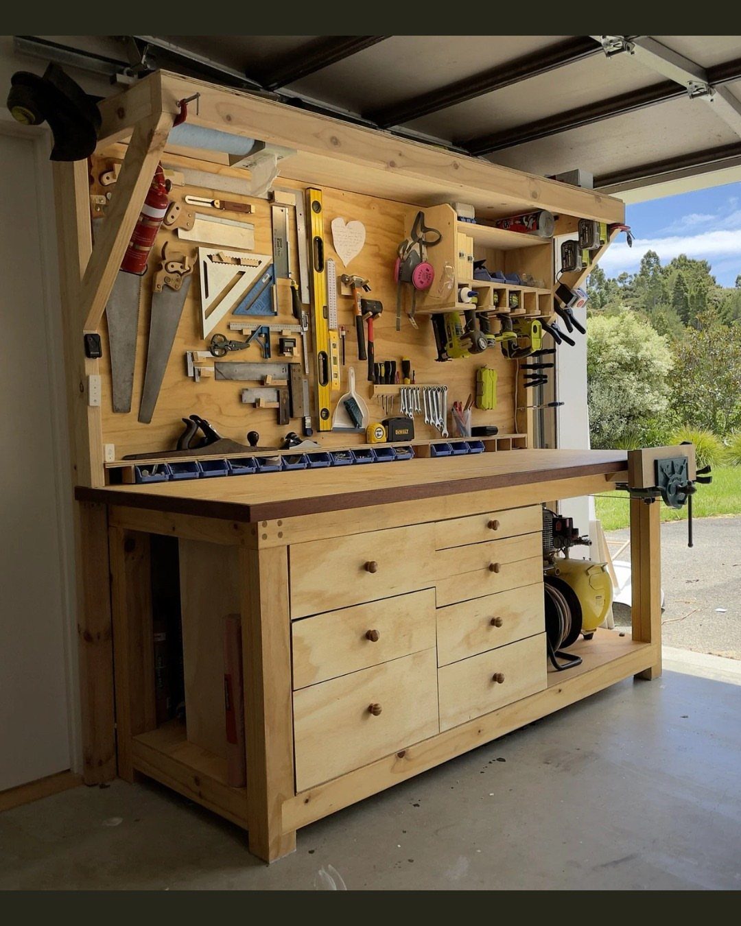 A wooden workbench with integrated drawers and a tool organizer wall filled with hand tools.