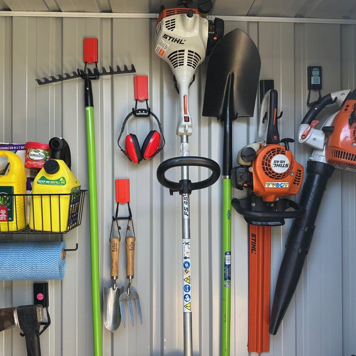 Garden tools like rakes, shovels, and trimmers hanging on a grey corrugated metal wall.