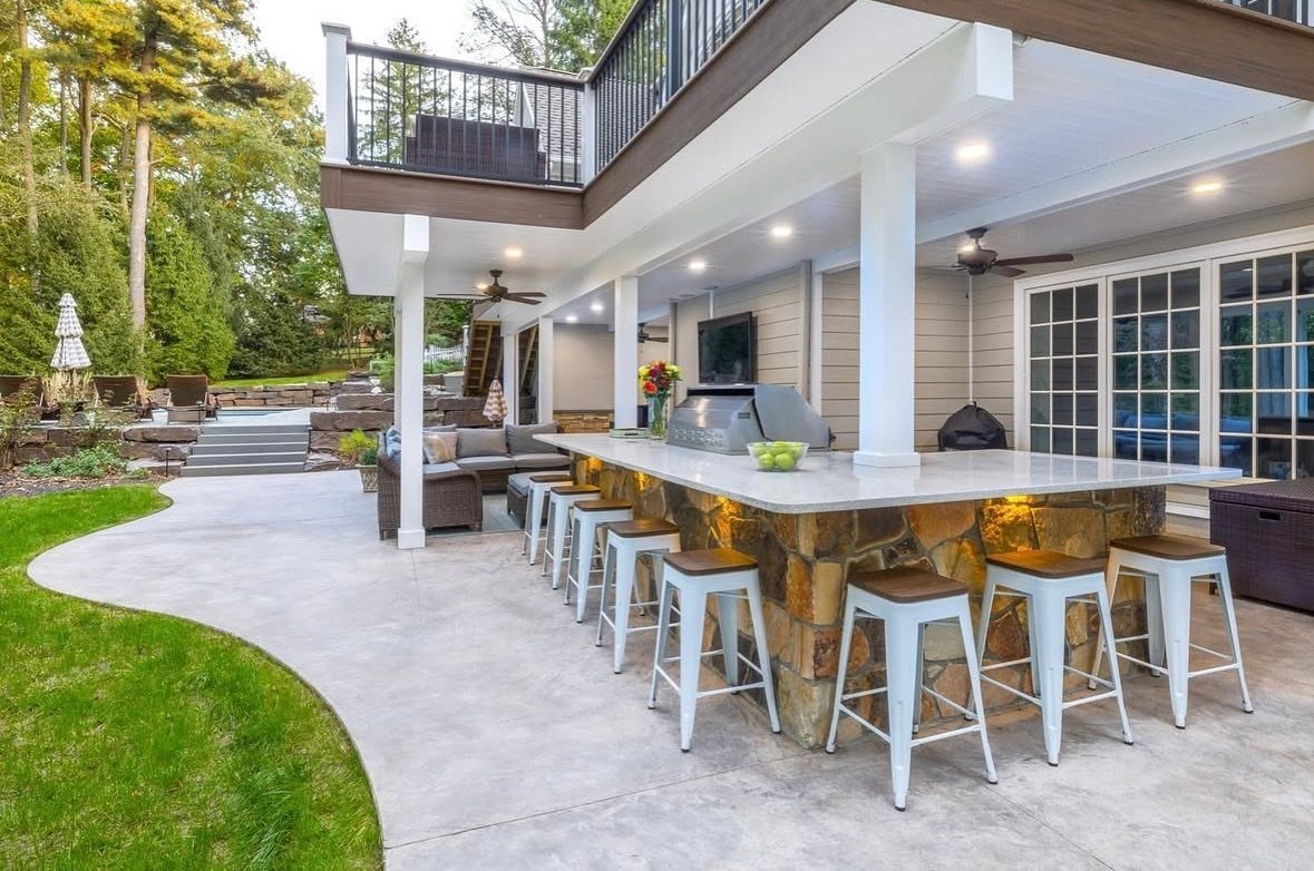 A stone outdoor bar with white stools and lounge seating situated under a large wooden deck.