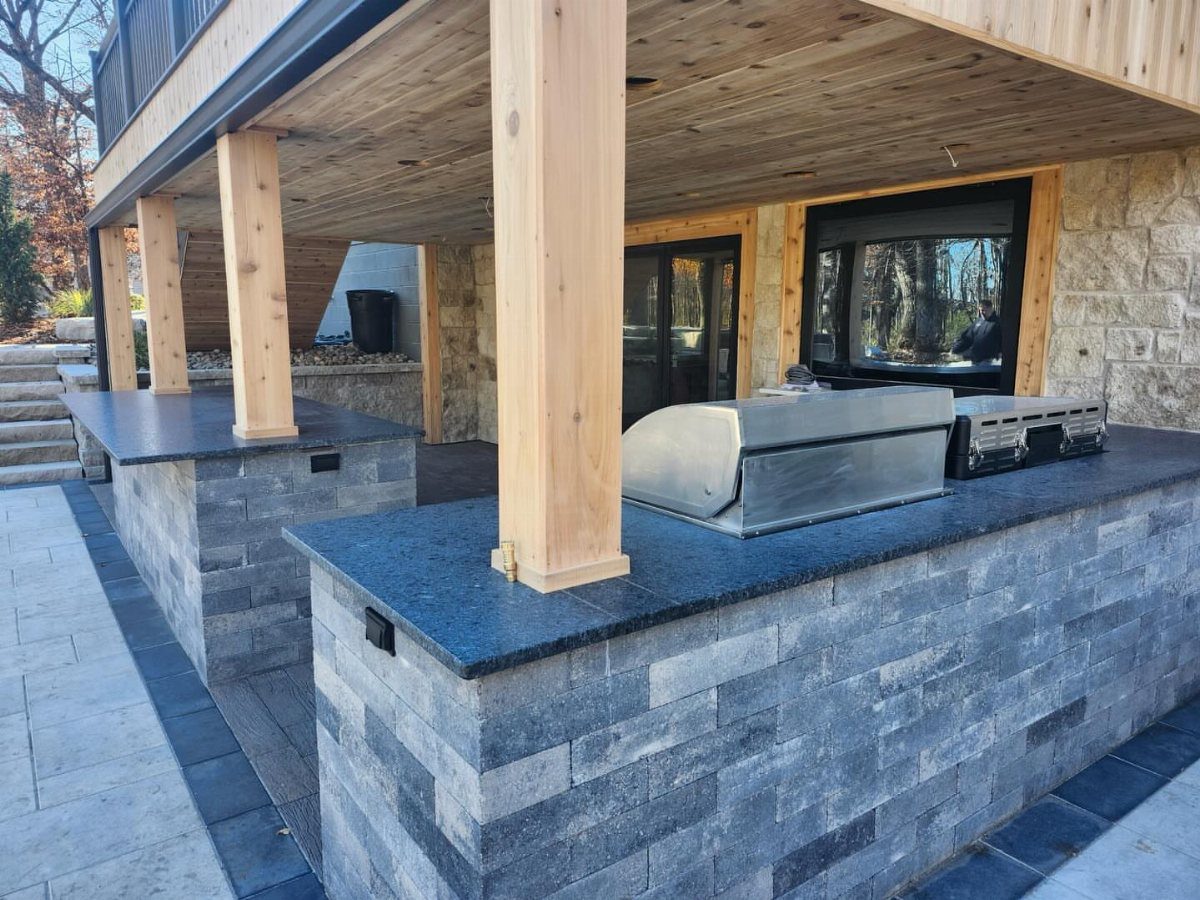 Outdoor kitchen with stone countertops and a stainless steel grill located under a wooden deck.