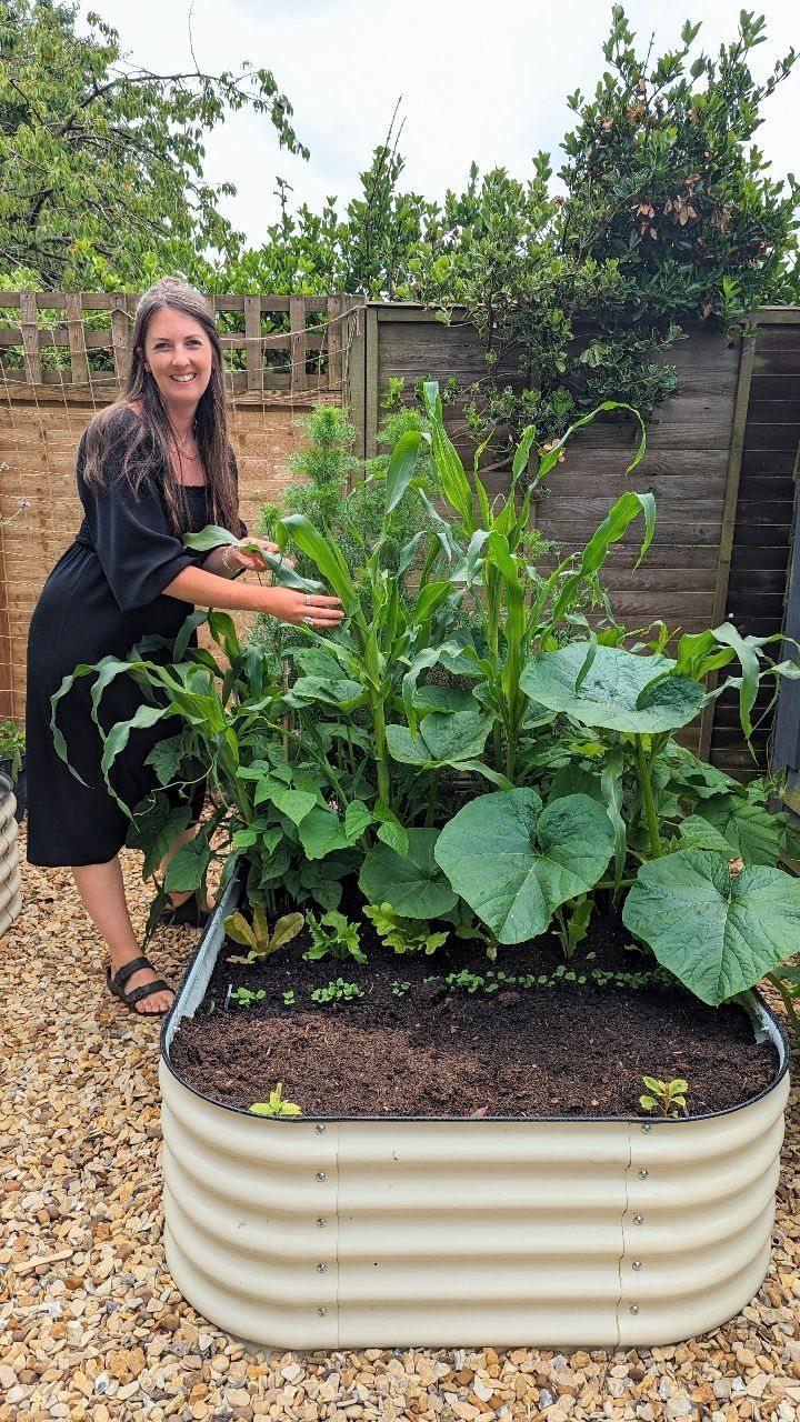 A woman smiling beside a raised garden bed containing tall corn and large squash leaves.