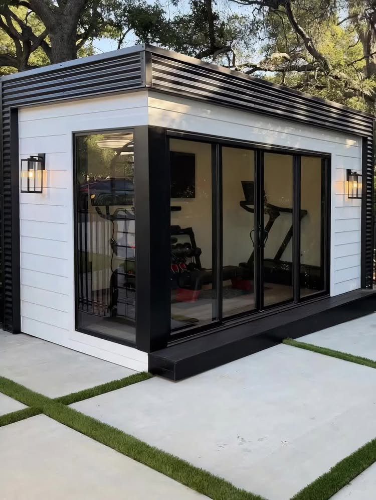 Modern white backyard gym shed with large glass doors revealing a treadmill and weights inside.