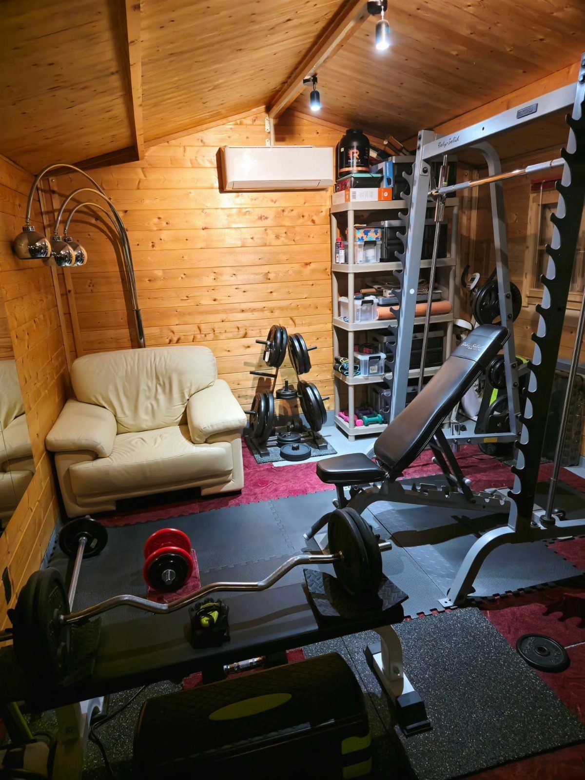 A wooden shed interior outfitted with weightlifting equipment, a bench, and a comfortable armchair.