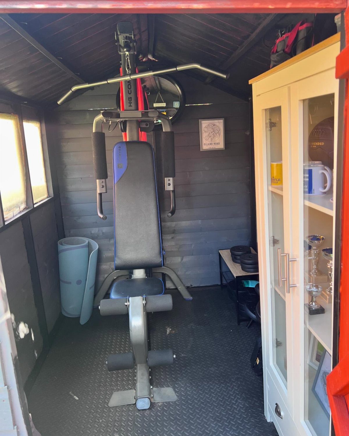 Weight machine positioned on black rubber mats inside a small wooden backyard gym shed.